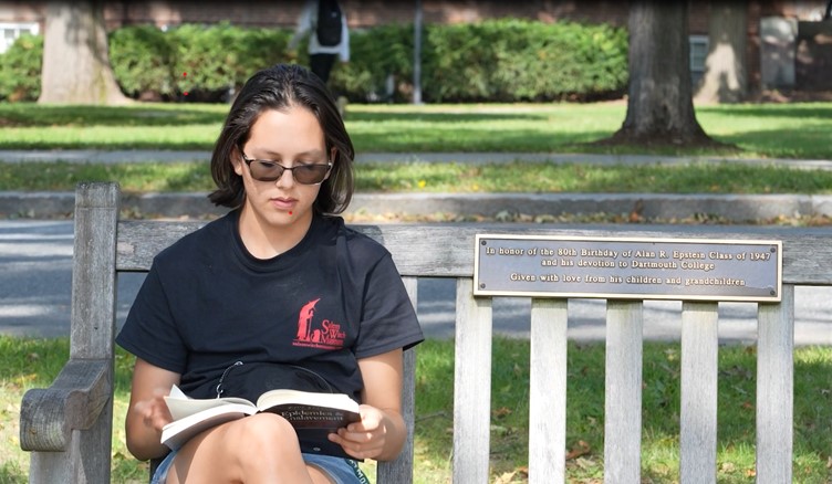 Student reading a book on a bench on the Dartmouth green.